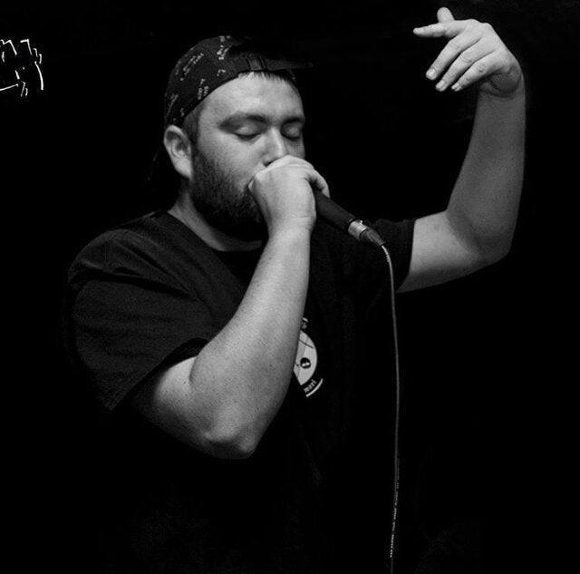 Black and white photo of Ash The Author performing live, wearing a black t-shirt and a patterned cap, passionately delivering lyrics into a microphone with one hand raised in expression.