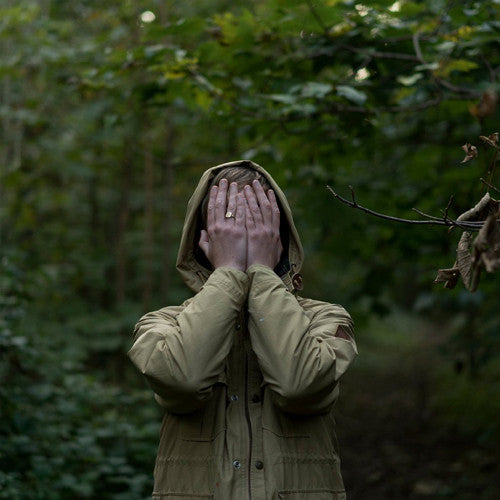Benedict standing in a lush green forest, wearing a hooded beige coat, covering their face with both hands. The image has a moody, introspective atmosphere, blending nature and anonymity.