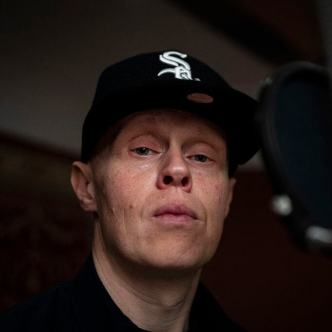 Cappo, UK-based rapper and lyricist, wearing a black White Sox cap and black hoodie, captured in a studio setting with a microphone in the foreground.