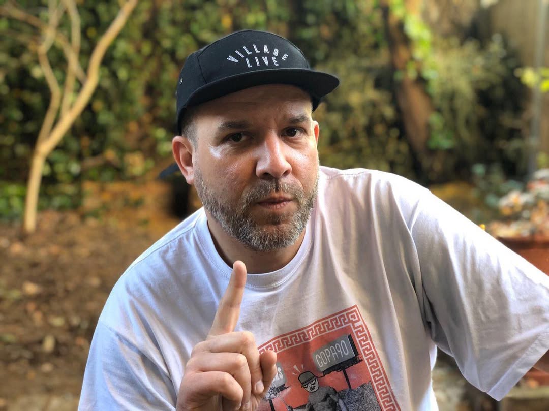 A man wearing a black Village Live five-panel cap and a white graphic T-shirt, pointing his finger while sitting outdoors.

