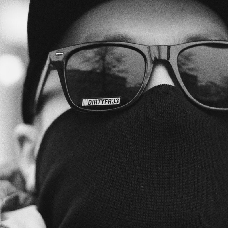 Black and white close-up portrait of DIRTYTHREE wearing sunglasses with "DIRTYFR33" branding on the frame and a black face covering, creating a mysterious and enigmatic look.