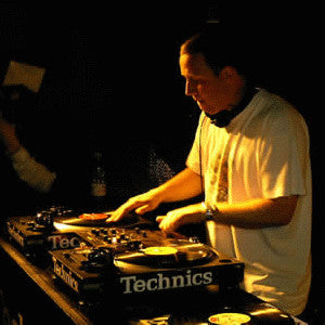 DJ Mr Thing performing live, standing behind a set of Technics turntables, wearing headphones around his neck and a white t-shirt, illuminated by warm stage lighting against a dark background.