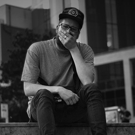 Black and white portrait of Ded Tebiase, sitting on outdoor steps in an urban setting. Wearing a cap and glasses, he rests his hand over his mouth in a relaxed pose, with a contemplative expression.