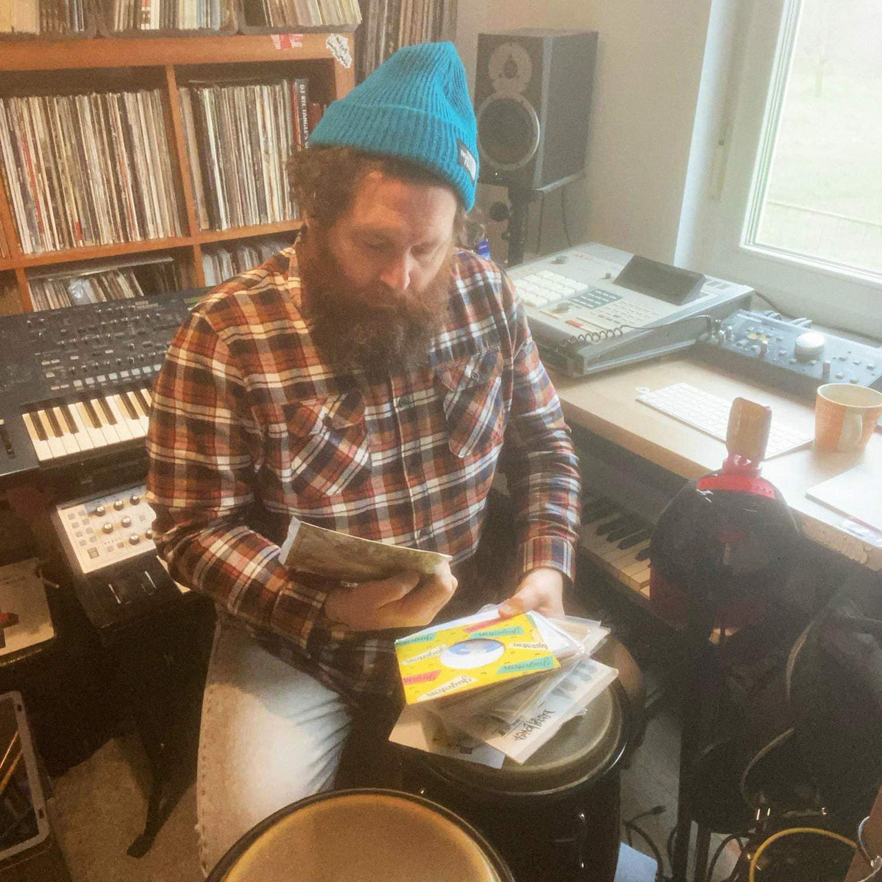 Dirty Hairy, UK-based DJ and producer, sitting in his home studio surrounded by vinyl records, samplers, and keyboards, wearing a blue beanie and flannel shirt while digging through a crate of records.