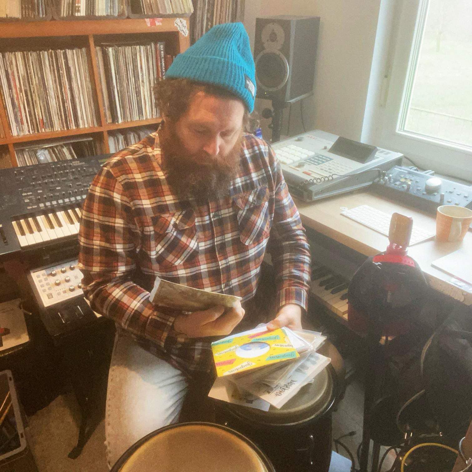 Dirty Hairy, UK-based DJ and producer, sitting in his home studio surrounded by vinyl records, samplers, and keyboards, wearing a blue beanie and flannel shirt while digging through a crate of records.