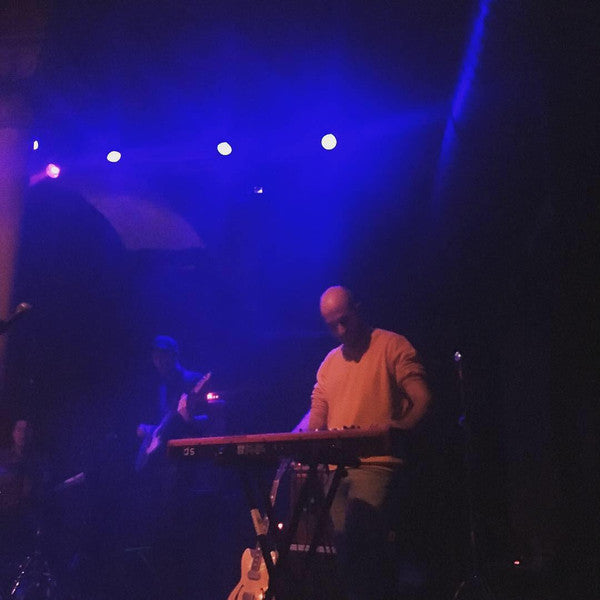 Doran Segal performing live on stage, playing the keyboard under atmospheric blue and purple stage lighting. A bassist is visible in the background.