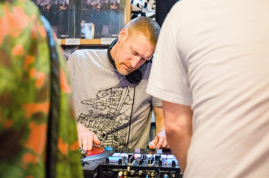 Evil Ed performing a DJ set, wearing headphones and a graphic t-shirt, focusing on the turntables.