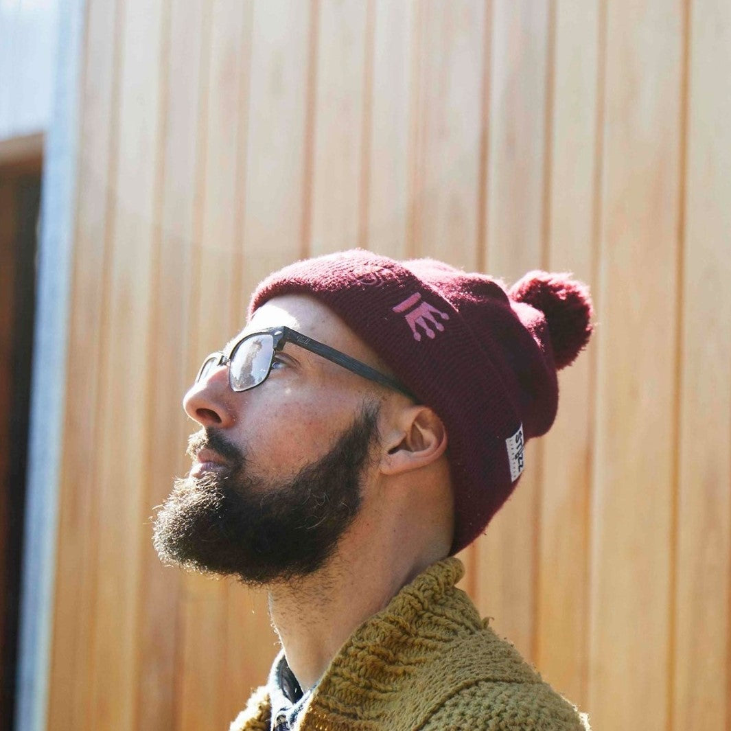 Gas-Lab wearing a maroon beanie and glasses, looking up against a wooden panel background.