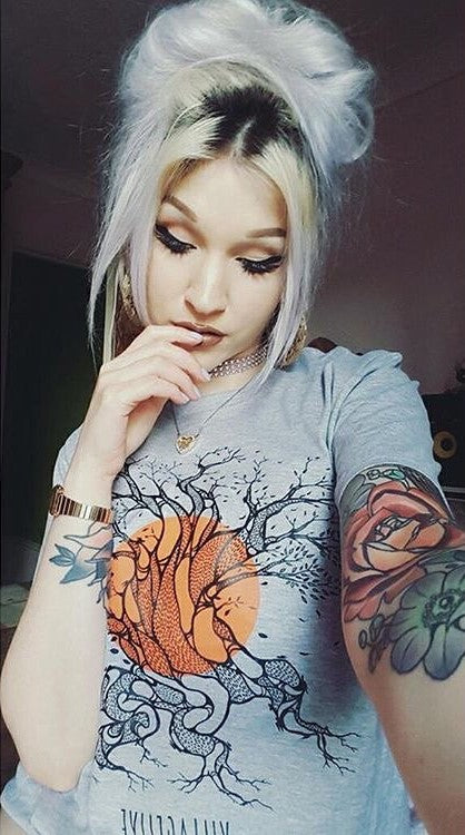 A woman with platinum blonde hair and tattoos wearing a Village Live graphic T-shirt, posing thoughtfully.