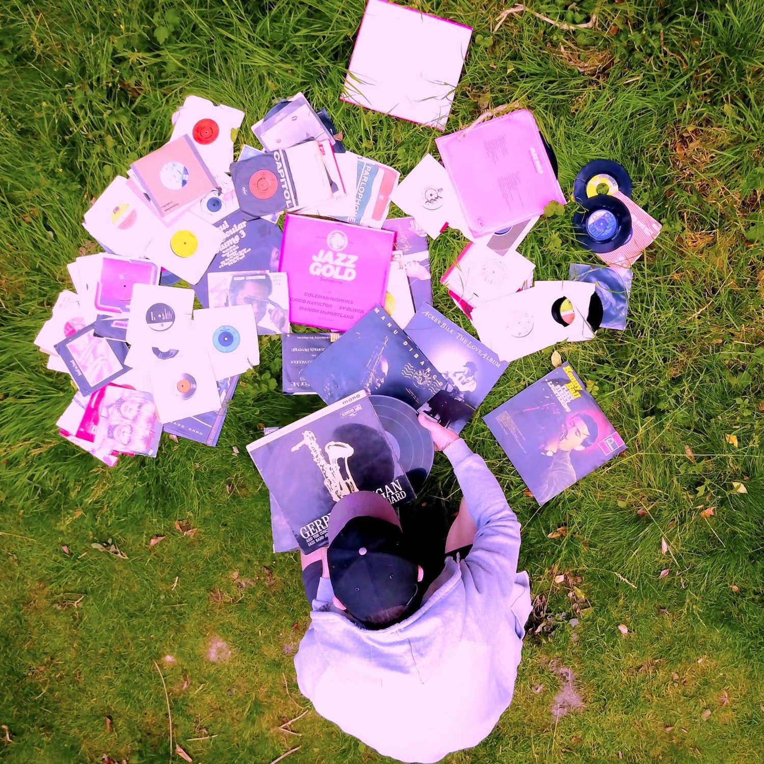 Isaintjames sitting on grass, surrounded by an array of purple and pink vinyl records and cassette tapes, engaged in selecting music.