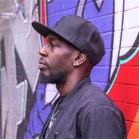 Justo The MC standing in front of a graffiti wall, wearing a black cap and jacket.