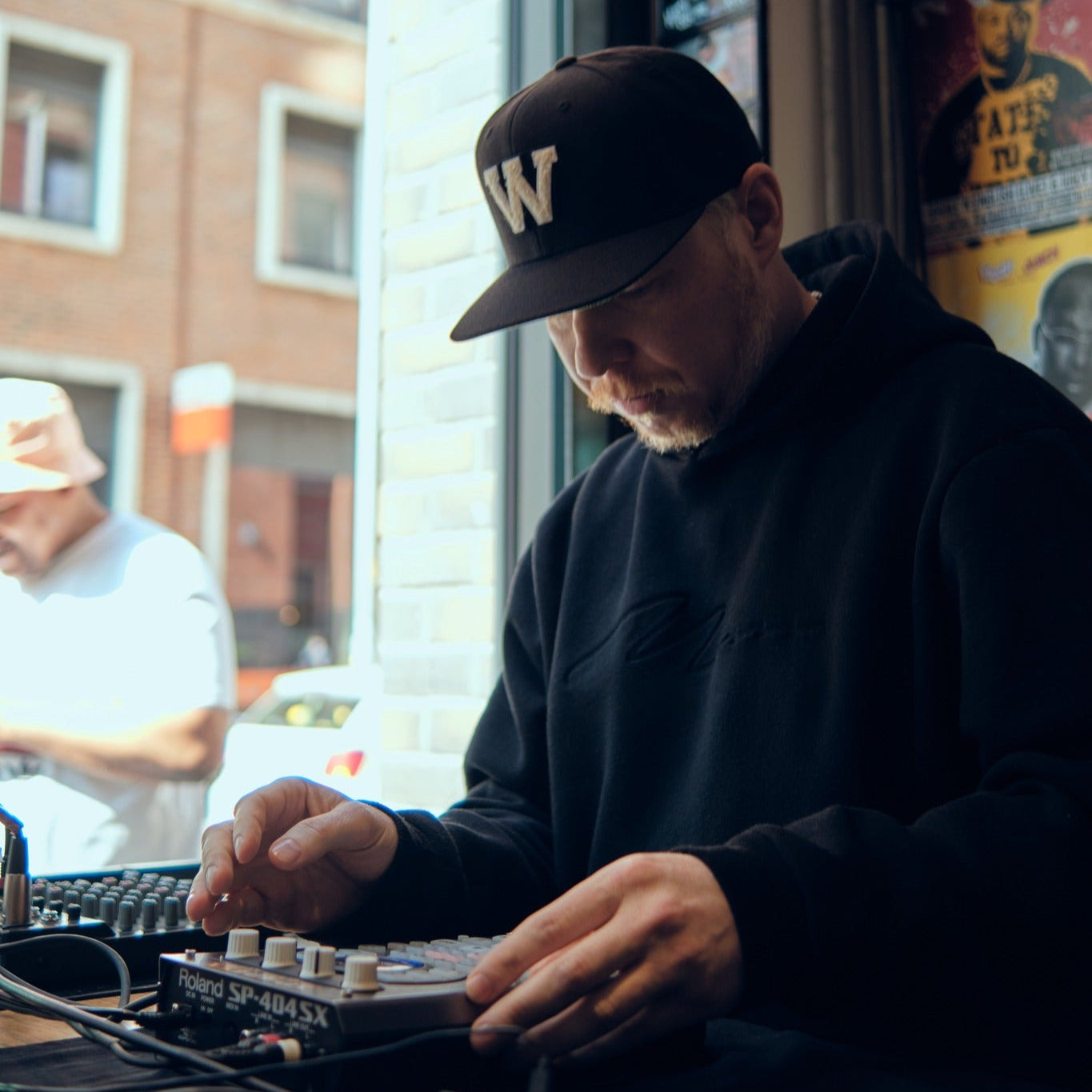 Kuartz performing live on an SP-404SX sampler, wearing a black hoodie and cap, with a focused expression in an urban setting.