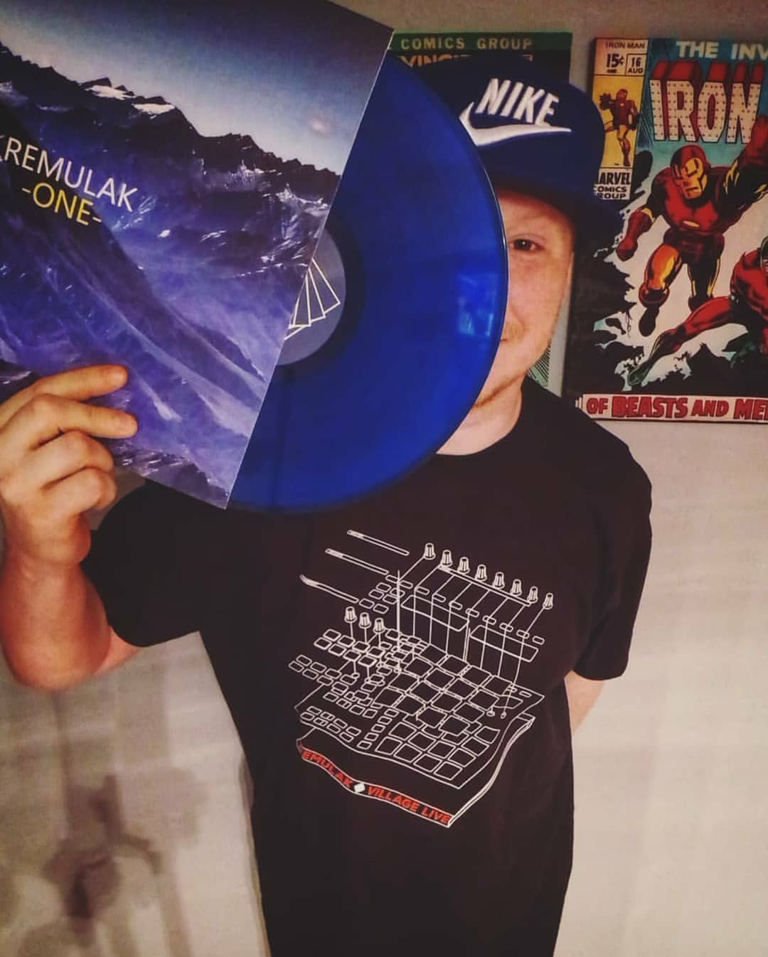 A man in a Nike cap holding up a blue vinyl record while wearing a black Village Live T-shirt with a chemistry-inspired design.
