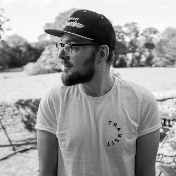 Black and white portrait of Mecca:83, a bearded man wearing a cap and glasses, looking to the side in an outdoor setting with trees in the background.