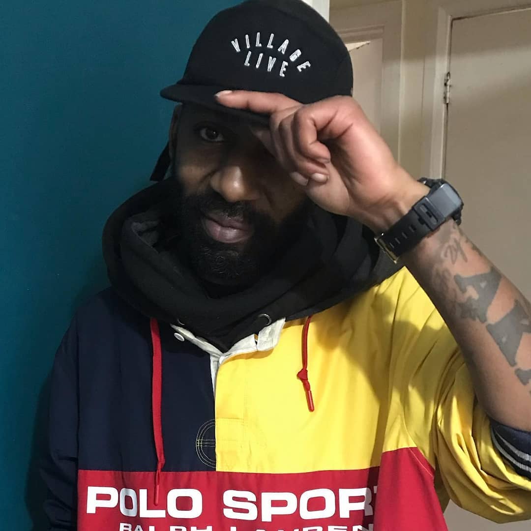 Parky, UK hip-hop artist, tipping his Village Live cap while wearing a vintage Polo Sport jacket, standing against a teal wall.