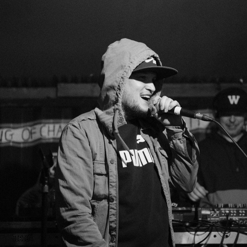 Black and white photo of Pique Roscoe performing live on stage, wearing a hooded jacket and cap while holding a microphone, delivering an energetic rap performance.