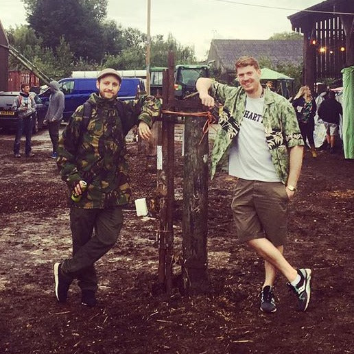 Remulak and Joey Deez standing outdoors at a muddy festival, dressed casually, leaning against a rustic wooden post.