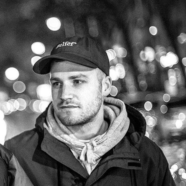 Slim Beats in a black-and-white portrait, wearing a cap and hoodie, standing against a blurred cityscape with glowing streetlights in the background. The image captures a thoughtful and urban aesthetic.