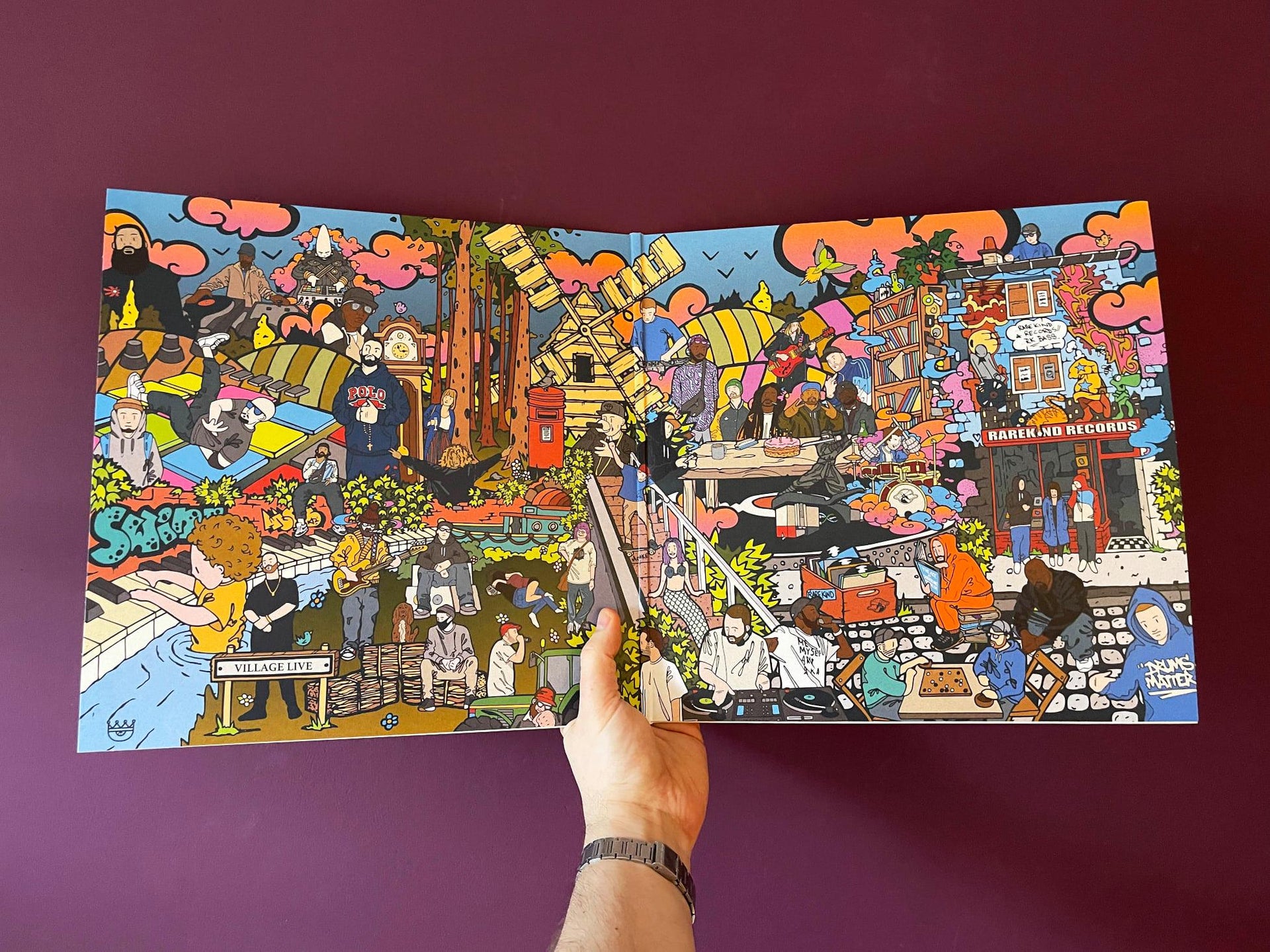A hand holding open the vibrant gatefold artwork of "Village Live Is Five," featuring a highly detailed, colorful illustration depicting a lively urban scene filled with musicians, record shops, and artistic elements.