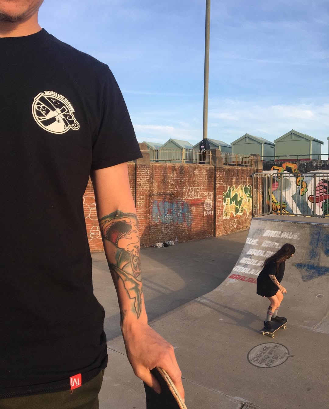 A person wearing a black Village Live OG T-shirt with a skateboarding scene in the background at a skatepark.