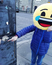 Person wearing a large emoji mask placing a Village Live Records sticker on a metal surface in an urban setting.